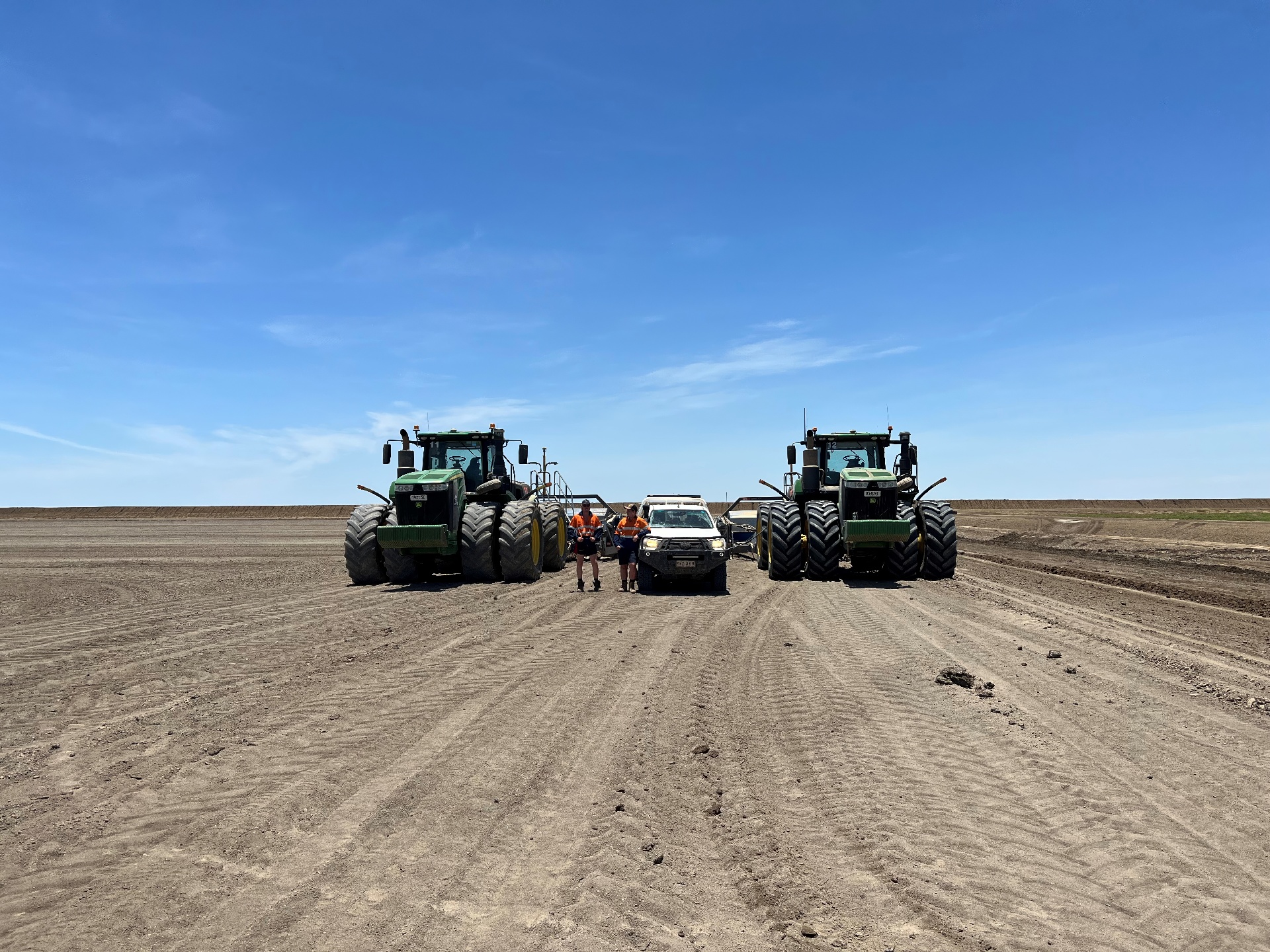 Two tractors are parked in a dirt field. In between them two people in hi-viz vests standing next to a white ute